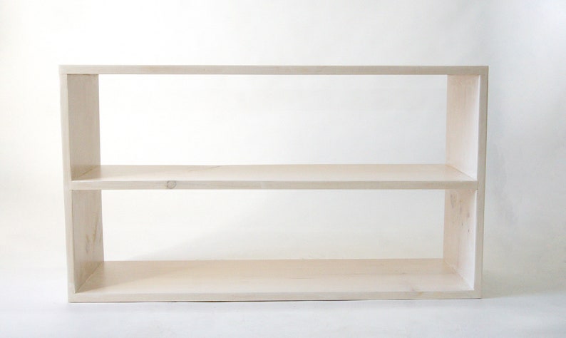 May include: A rectangular, light-colored wooden shelf unit featuring two horizontal tiers. The structure is composed of a top panel, a bottom panel, two side panels, and a single middle shelf, creating two open storage compartments with a smooth finish.