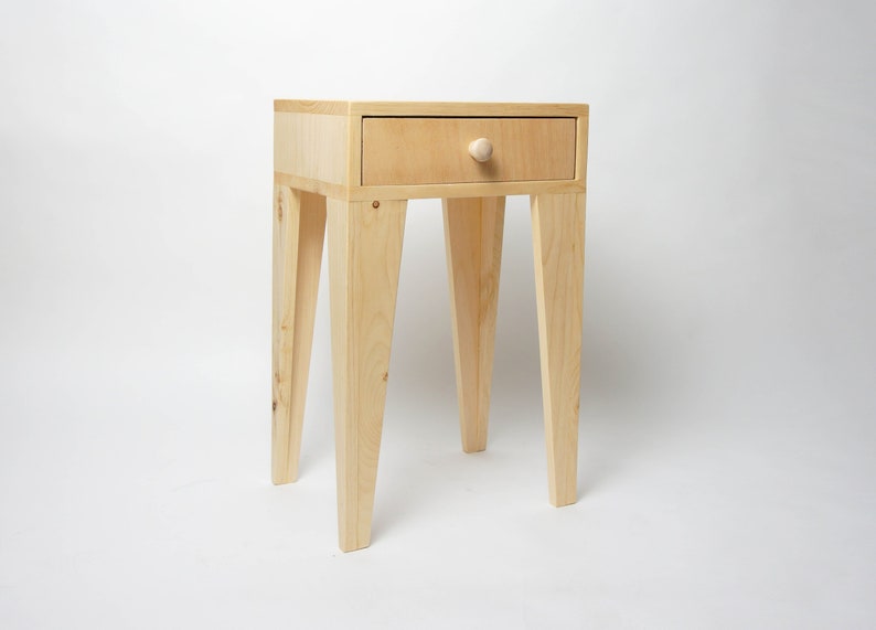 May include: A light-coloured wooden bedside table with a single drawer and a round knob. The table has four angled legs and is made of light-coloured wood. The drawer is centred on the front of the table.