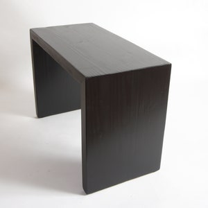 May include: A rectangular bench with a dark brown, almost black, wood grain finish. The piece features a simple, continuous U-shaped design with a flat top surface and two vertical side panels that serve as legs, showcasing a smooth, solid construction.