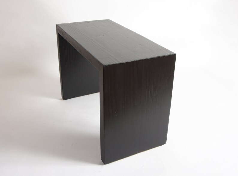 May include: A rectangular bench with a dark brown, almost black, wood grain finish. The piece features a simple, continuous U-shaped design with a flat top surface and two vertical side panels that serve as legs, showcasing a smooth, solid construction.