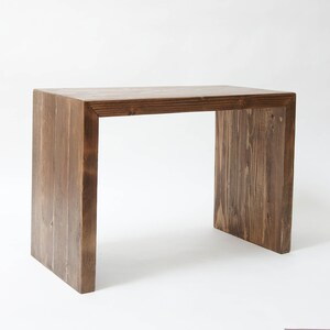 May include: A rectangular wooden desk with a dark brown stained finish and a visible wood grain pattern. The desk features a waterfall design where the flat top surface connects directly to two vertical side panels, creating a continuous structure.