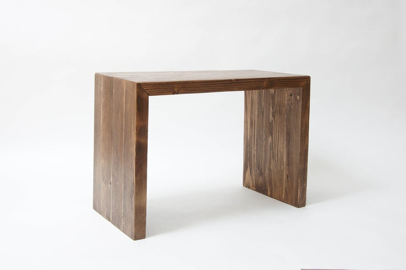 May include: A rectangular wooden desk with a dark brown stained finish and a visible wood grain pattern. The desk features a waterfall design where the flat top surface connects directly to two vertical side panels, creating a continuous structure.