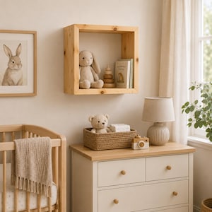 May include: A square light-colored wooden wall shelf containing a plush rabbit, a wooden stacking toy, and a book. The shelf is mounted above a white dresser with four drawers, which holds a woven basket with a teddy bear and a table lamp.