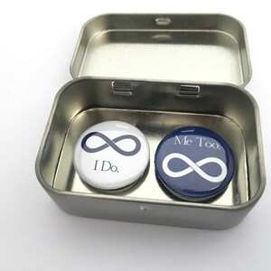 May include: A small metal tin box with two round buttons inside. The buttons are white and blue with infinity symbols and the text "I Do." and "Me Too."