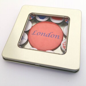 Puede incluir: Una lata de metal plateada con una ventana de pl&aacute;stico transparente que muestra una colecci&oacute;n de botones redondos. Los botones son blancos con dise&ntilde;os azules y rojos. Un bot&oacute;n es rojo con la palabra "London" en letras azules.