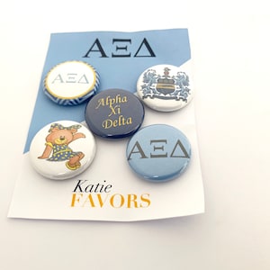 May include: Five round buttons with different designs. The buttons are white, blue, and navy blue. The designs include the letters "ΑΕΔ", a crest, a teddy bear, and the words "Alpha Xi Delta". The buttons are on a light blue background with the words "Katie FAVORS" in yellow.