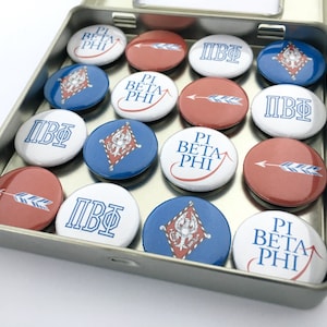 May include: A tin box filled with round, colorful buttons. The buttons feature the Greek letters "ΠΒΦ" and the words "Pi Beta Phi" in blue and red lettering. Some buttons have an arrow design.