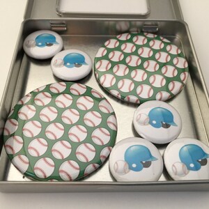 May include: A metal tin with eight round baseball-themed buttons. The buttons feature a green and white baseball pattern, a blue baseball helmet with a white baseball, and a white background with a blue baseball helmet and a white baseball.