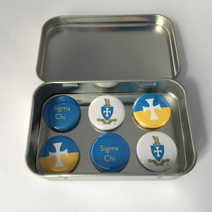 Puede incluir: Una lata de metal plateada con seis botones redondos azules y amarillos. Los botones tienen una cruz blanca, un escudo con una cruz y el texto "Sigma Chi".