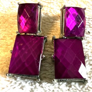 May include: A pair of drop earrings featuring two stacked, faceted, rectangular magenta stones set in silver-toned metal. Each earring consists of a smaller square-like top stone and a larger rectangular bottom stone with a geometric cut.