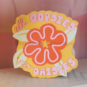 May include: A yellow sign with the words "all oopsies no daisies" in pink lettering. The sign features a large orange flower with pink outlines and green leaves. The sign is on a white surface.