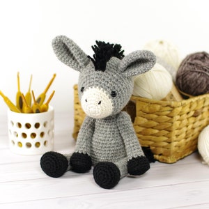 Puede incluir: Un juguete de burro de crochet gris con pezuñas negras y una crin negra. El burro está sentado sobre una superficie de madera blanca con una cesta de hilo en el fondo.