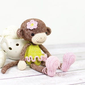 Puede incluir: Una muñeca de mono de crochet que lleva un vestido amarillo con ribetes rosas y una flor rosa en la cabeza. El mono está sentado sobre una superficie de madera blanca.