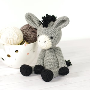 May include: A grey crocheted donkey toy with black hooves and a black mane. The donkey is sitting on a white wooden surface with balls of yarn in a white ceramic bowl.