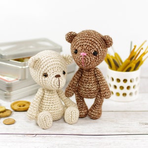 May include: Two crocheted teddy bears, one brown and one cream, sitting on a white wooden surface. The brown bear has a pink nose and the cream bear has brown eyes.
