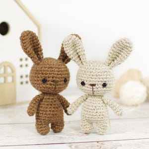 Könnte beinhalten: Zwei gehäkelte Hasen-Plüschtiere, eines braun und eines beige, stehen nebeneinander auf einer weißen Holzoberfläche. Beide Hasen haben schwarze Augen und braune Nasen.