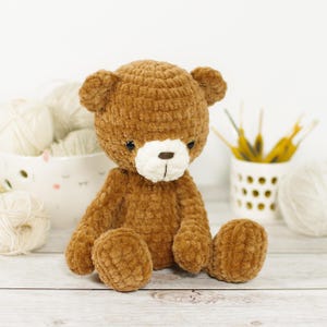 Könnte beinhalten: Ein handgefertigter brauner Teddybär mit weißer Schnauze und schwarzen Augen. Das Plüschtier sitzt mit ausgestreckten Beinen. Der Bär ist aus weichem, strukturiertem Garn gefertigt. Bastelbedarf befindet sich im Hintergrund.