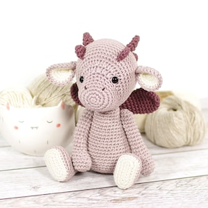 Peut inclure: Un dragon en crochet fait à la main, en fil rose clair avec des détails rose foncé. Le dragon a de petites ailes, des cornes et des accents blancs sur les oreilles et les pieds. Il est posé sur une surface en bois blanc, avec de la laine et un bol décoratif en arrière-plan.