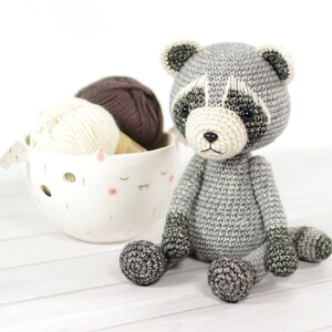 May include: A gray crocheted raccoon amigurumi toy sitting on a white wooden surface. The toy has black eyes and a black nose. There is a white ceramic bowl with two balls of yarn in the background.