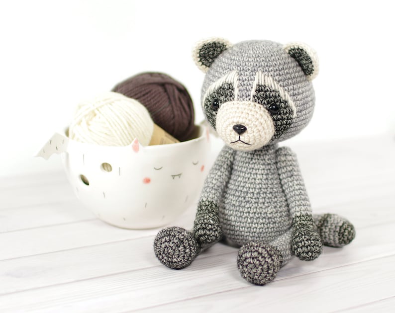 May include: A gray crocheted raccoon amigurumi toy sitting on a white wooden surface. The toy has black eyes and a black nose. There is a white ceramic bowl with two balls of yarn in the background.