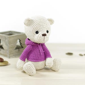 K&ouml;nnte beinhalten: Ein handgeh&auml;kelter Teddyb&auml;r mit cremefarbenem K&ouml;rper und lila Kapuzenpullover. Der B&auml;r hat schwarze Augen und eine braune Nase und sitzt auf einer hellen Oberfl&auml;che. Ein bezauberndes Geschenk f&uuml;r Kinder. H&ouml;he ca. 20 cm.