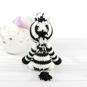 K&ouml;nnte beinhalten: Ein schwarz-wei&szlig; gestreiftes geh&auml;keltes Zebra-Spielzeug mit einem flauschigen Schwanz. Das Spielzeug sitzt auf einer wei&szlig;en Holzoberfl&auml;che.