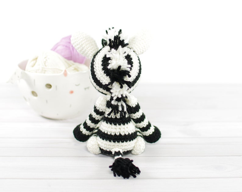 K&ouml;nnte beinhalten: Ein schwarz-wei&szlig; gestreiftes geh&auml;keltes Zebra-Spielzeug mit einem flauschigen Schwanz. Das Spielzeug sitzt auf einer wei&szlig;en Holzoberfl&auml;che.