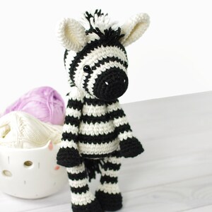 K&ouml;nnte beinhalten: Ein gestricktes Zebra-Spielzeug mit schwarzen und wei&szlig;en Streifen, schwarzen Augen und einer schwarzen Nase. Das Zebra steht auf einer wei&szlig;en Holzoberfl&auml;che.
