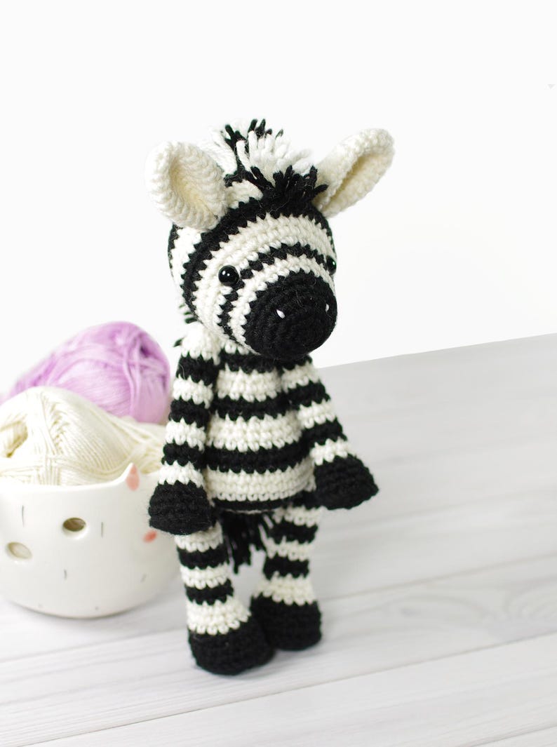 K&ouml;nnte beinhalten: Ein gestricktes Zebra-Spielzeug mit schwarzen und wei&szlig;en Streifen, schwarzen Augen und einer schwarzen Nase. Das Zebra steht auf einer wei&szlig;en Holzoberfl&auml;che.