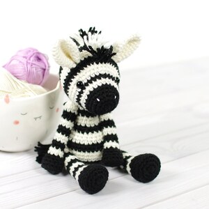 K&ouml;nnte beinhalten: Ein gestricktes Zebra-Spielzeug in Schwarz-Wei&szlig; auf einer wei&szlig;en Holzoberfl&auml;che. Das Zebra hat schwarze Augen und eine schwarze Nase. Im Hintergrund befinden sich zwei Wollkn&auml;uel.