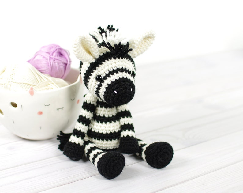 K&ouml;nnte beinhalten: Ein gestricktes Zebra-Spielzeug in Schwarz-Wei&szlig; auf einer wei&szlig;en Holzoberfl&auml;che. Das Zebra hat schwarze Augen und eine schwarze Nase. Im Hintergrund befinden sich zwei Wollkn&auml;uel.