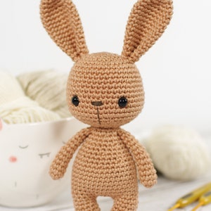 Amigurumi bunny, standing in front of a yarn bowl , crochet hooks and balls of yarn