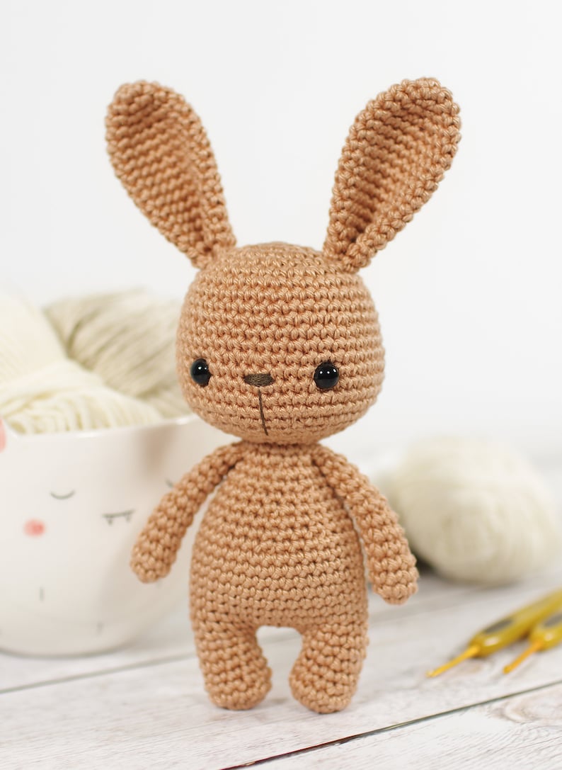 Amigurumi bunny, standing in front of a yarn bowl , crochet hooks and balls of yarn