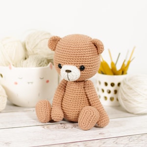 Op de afbeelding: Een gehaakte teddybeer in een zittende positie. De beer is lichtbruin met een witte snuit en zwarte ogen. De achtergrond bevat garen en knutselspullen, wat duidt op een handgemaakt item.