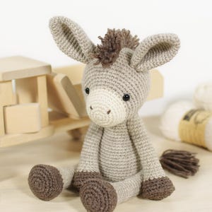 May include: A crocheted donkey soft toy featuring a light grey body, a cream-coloured muzzle, dark brown hooves, and a dark brown mane and tail. The donkey has two large, upright ears and small black eyes, sitting in an upright position.