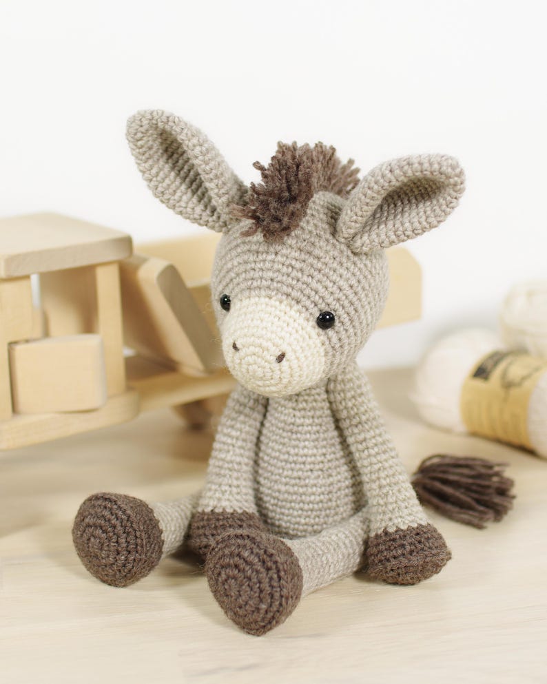 May include: A crocheted donkey soft toy featuring a light grey body, a cream-coloured muzzle, dark brown hooves, and a dark brown mane and tail. The donkey has two large, upright ears and small black eyes, sitting in an upright position.