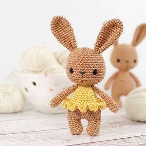 Small crocheted amigurumi bunny in a yellow ballerina outfit, standing in front of a yarn bowl and balls of yarn