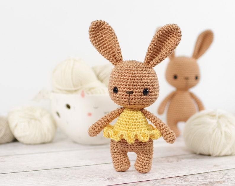 Small crocheted amigurumi bunny in a yellow ballerina outfit, standing in front of a yarn bowl and balls of yarn