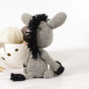 May include: A handmade crochet donkey in shades of gray with black accents. The donkey has a black mane and tail, and is sitting down. A white ceramic bowl filled with yarn and crochet hooks are in the background.