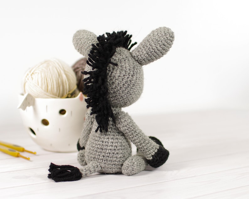 May include: A handmade crochet donkey in shades of gray with black accents. The donkey has a black mane and tail, and is sitting down. A white ceramic bowl filled with yarn and crochet hooks are in the background.