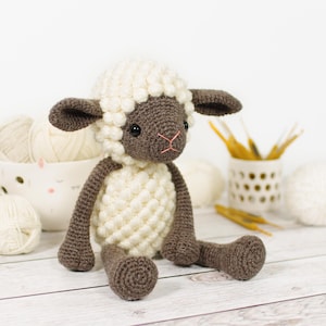 Puede incluir: Una muñeca de cordero de ganchillo hecha a mano con cuerpo, cabeza y orejas blancos y detalles marrones. El cordero tiene ojos negros y una boca cosida rosa. La muñeca está sentada sobre una superficie de madera blanca.