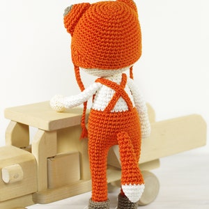 May include: A crocheted fox doll wearing orange overalls and a matching hat with white accents. The doll is standing next to a wooden toy truck.