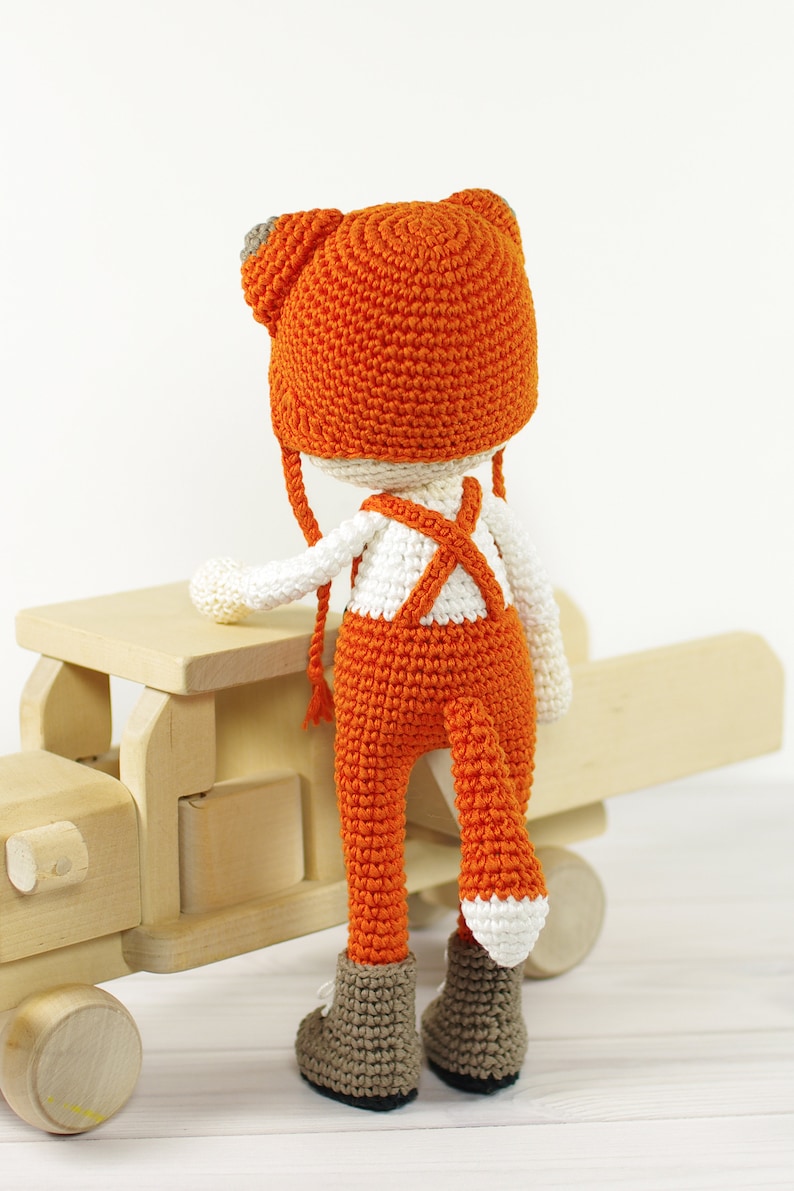 May include: A crocheted fox doll wearing orange overalls and a matching hat with white accents. The doll is standing next to a wooden toy truck.