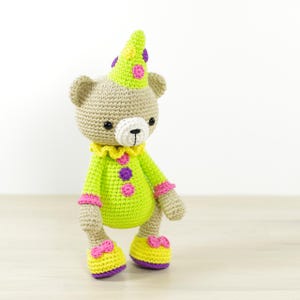 May include: A crocheted stuffed bear wearing a green and yellow clown costume with a purple and yellow hat. The bear has a white face with black eyes and a black nose.
