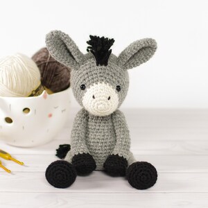 May include: A handmade crochet donkey toy in gray, white, and black. The donkey has large ears, a white muzzle, and black hooves. A white bowl with yarn and two yellow crochet hooks are in the background.
