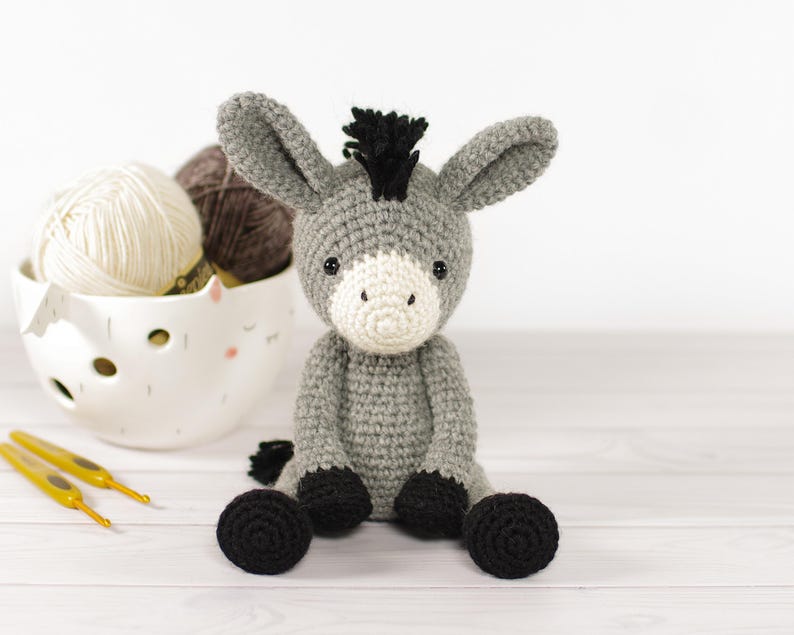 May include: A handmade crochet donkey toy in gray, white, and black. The donkey has large ears, a white muzzle, and black hooves. A white bowl with yarn and two yellow crochet hooks are in the background.