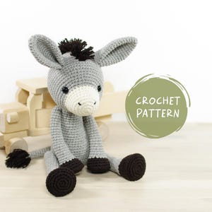 May include: A crocheted gray donkey toy with black hooves and a black mane. The donkey is sitting on a wooden toy truck. The text "CROCHET PATTERN" is on a green circle.