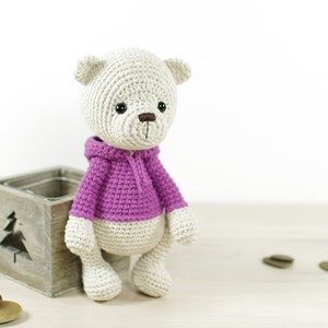May include: A crocheted teddy bear wearing a purple hooded sweater. The bear is sitting on a wooden box with a cutout of a tree.