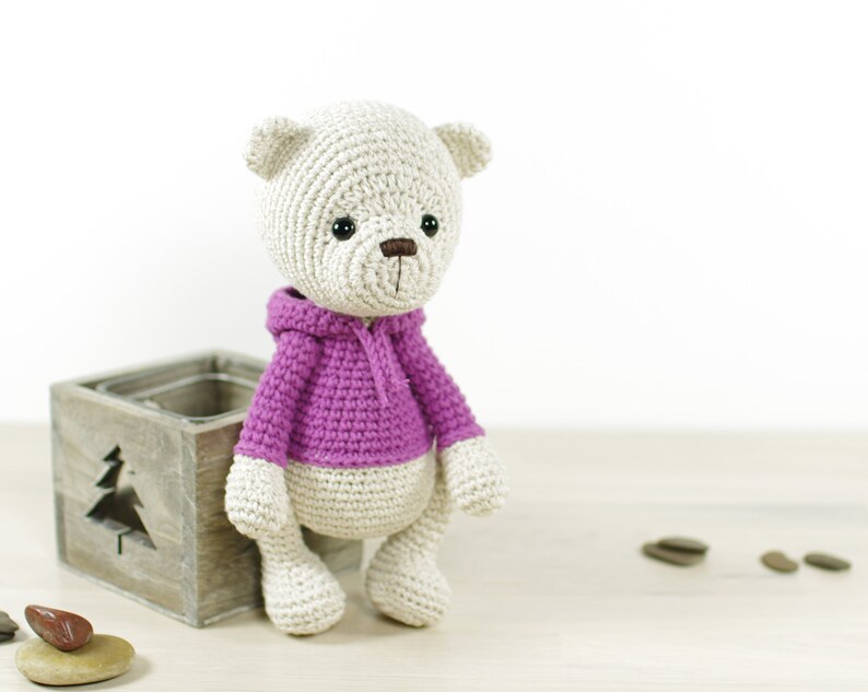 May include: A crocheted teddy bear wearing a purple hooded sweater. The bear is sitting on a wooden box with a cutout of a tree.