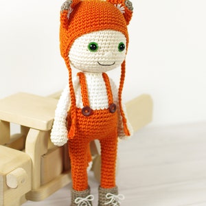 May include: A crocheted fox doll wearing orange overalls and a matching hat with fox ears. The doll has green eyes and a white face. The doll is standing next to a wooden toy.